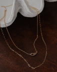 two 14 karat gold paperclip necklaces draped on a shell with dark wood tabletop