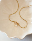 Gold snake chain necklace with freshwater pearl charm displayed in a cream ceramic dish by Sincerely Chain Co.
