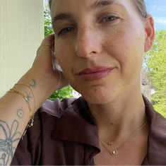 Woman with tattoos and jewelry outdoors, wearing a brown top.