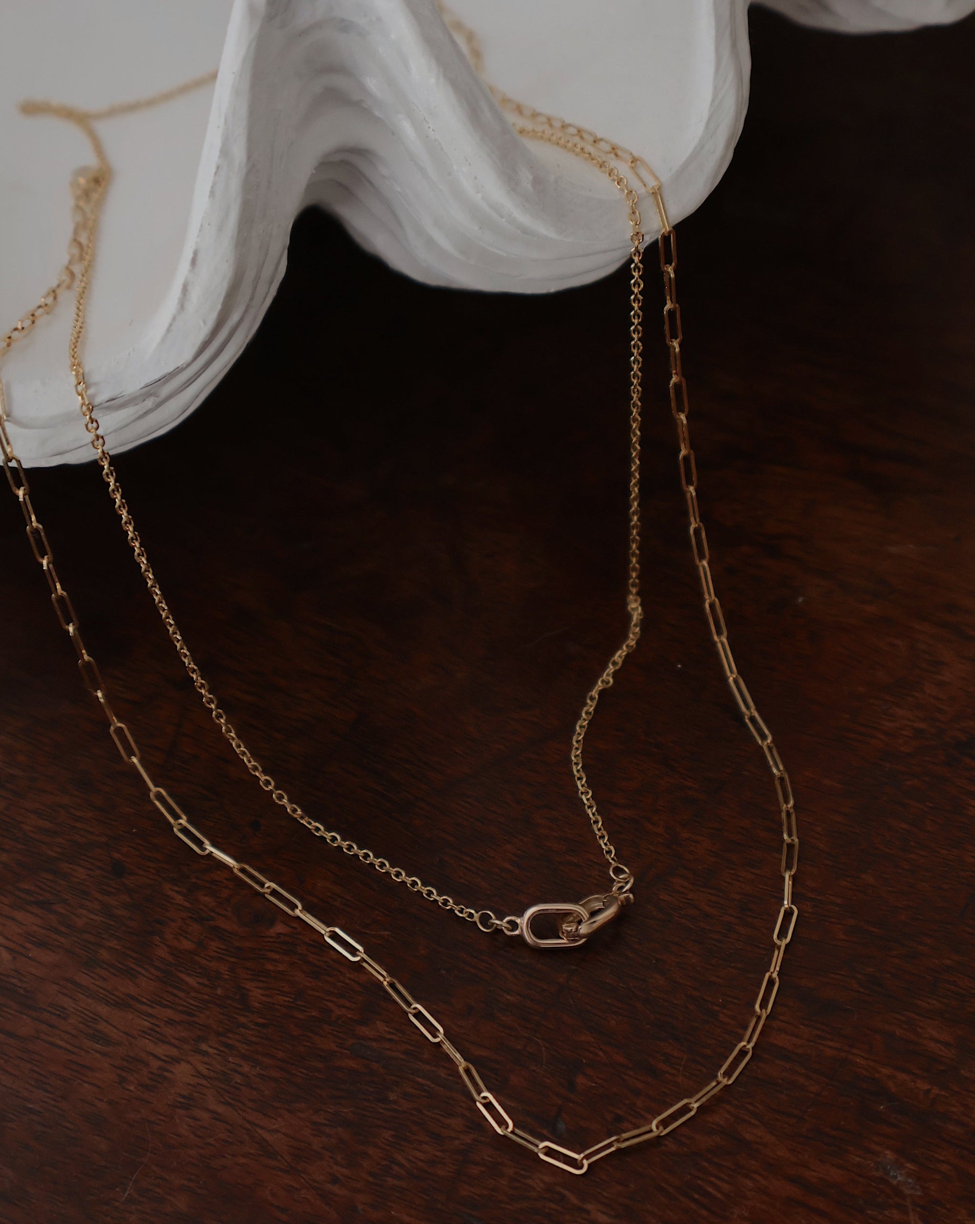 two 14 karat gold paperclip necklaces draped on a shell with dark wood tabletop