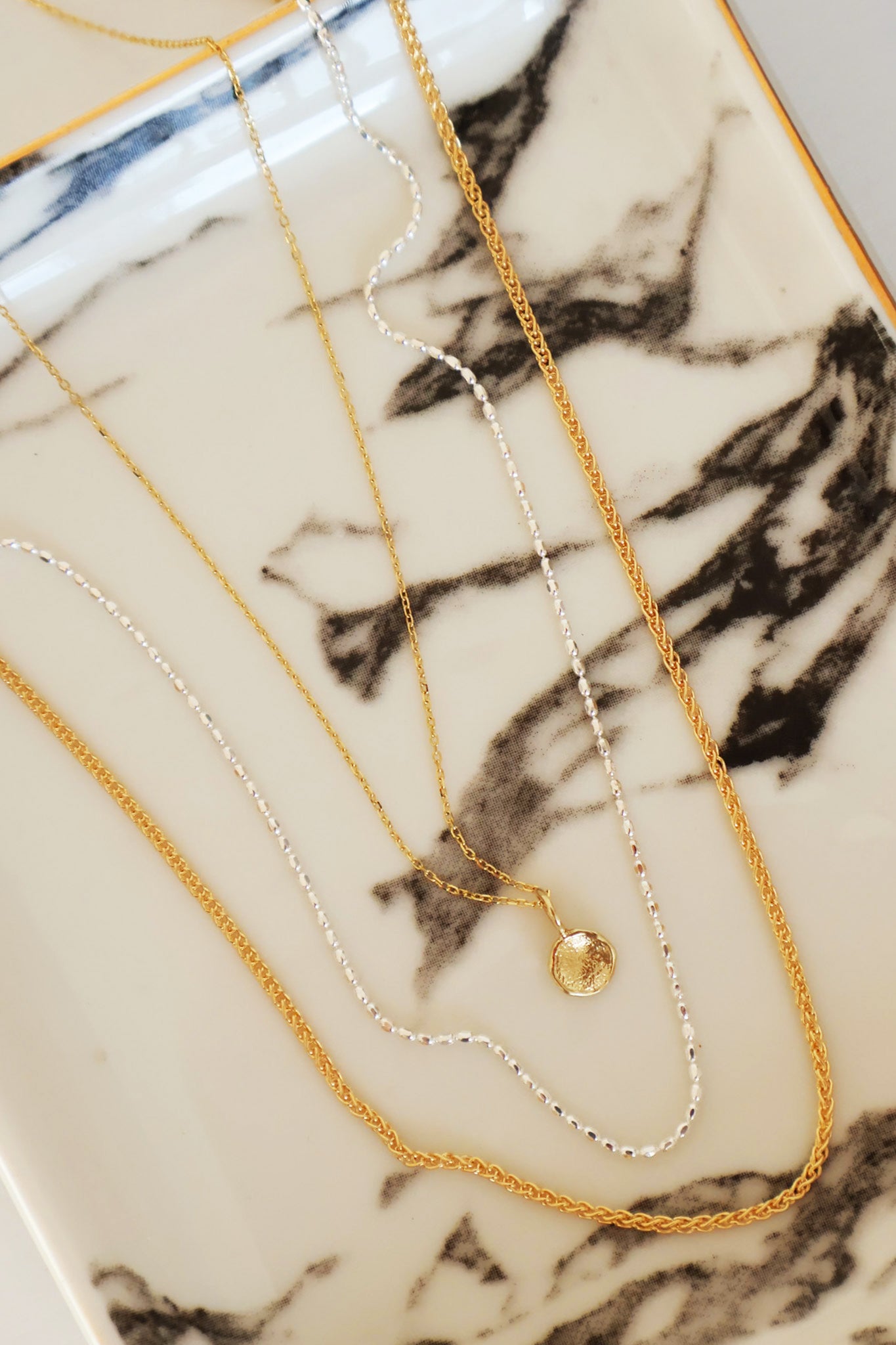 A selection of layered necklaces by Sincerely Chain Co. featuring a solid gold chain, sterling silver chain, and a gold chain with a mini disc charm, displayed on a marble surface.