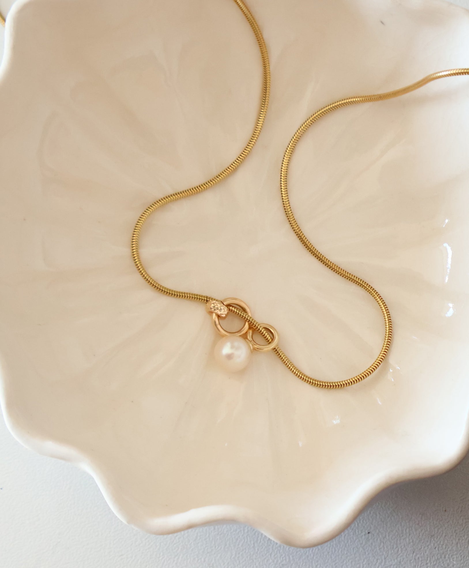 Gold snake chain necklace with freshwater pearl charm displayed in a cream ceramic dish by Sincerely Chain Co.