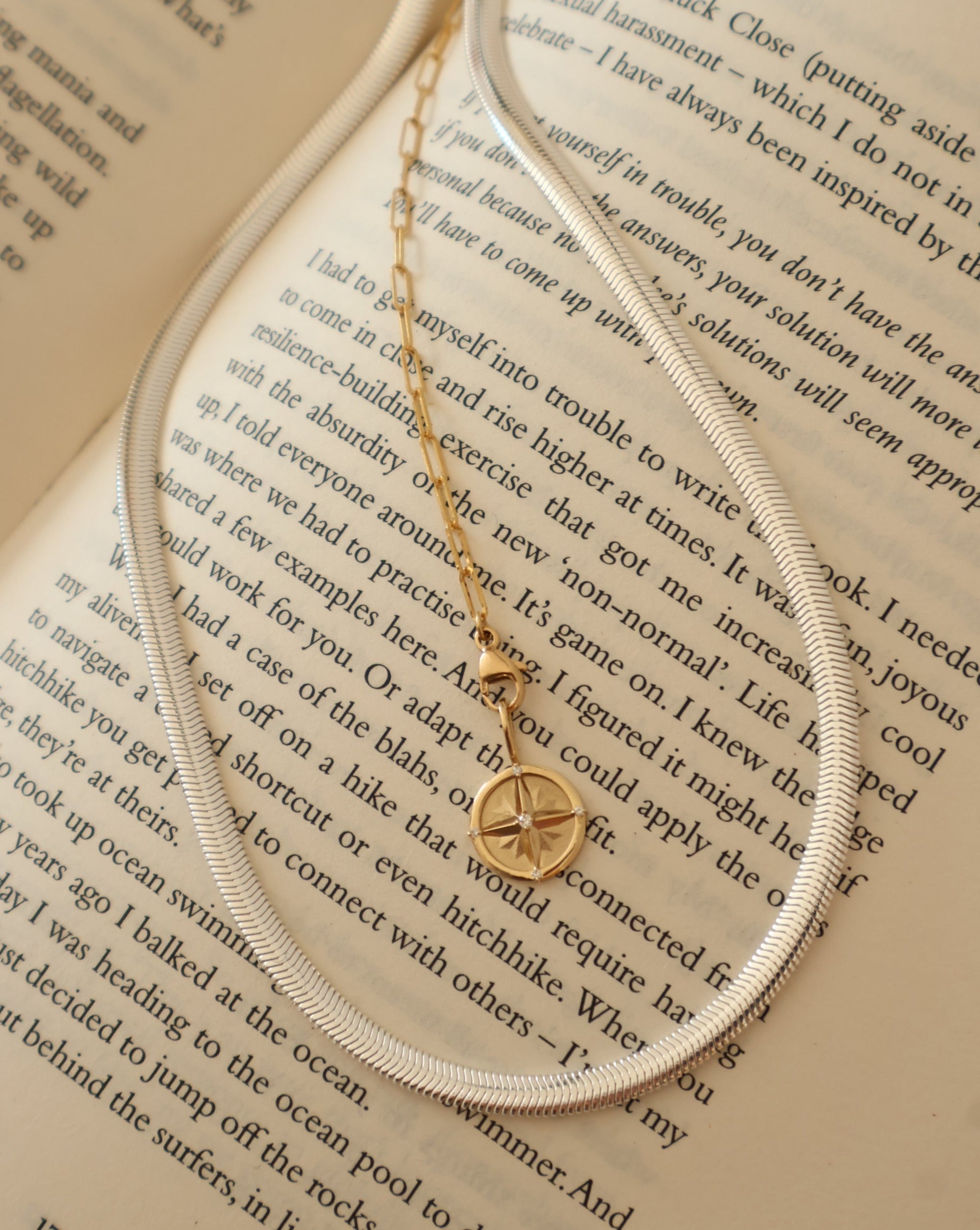 Sterling silver herringbone necklace and 14k gold compass pendant draped on an open book, fine jewelry by Sincerely Chain Co.